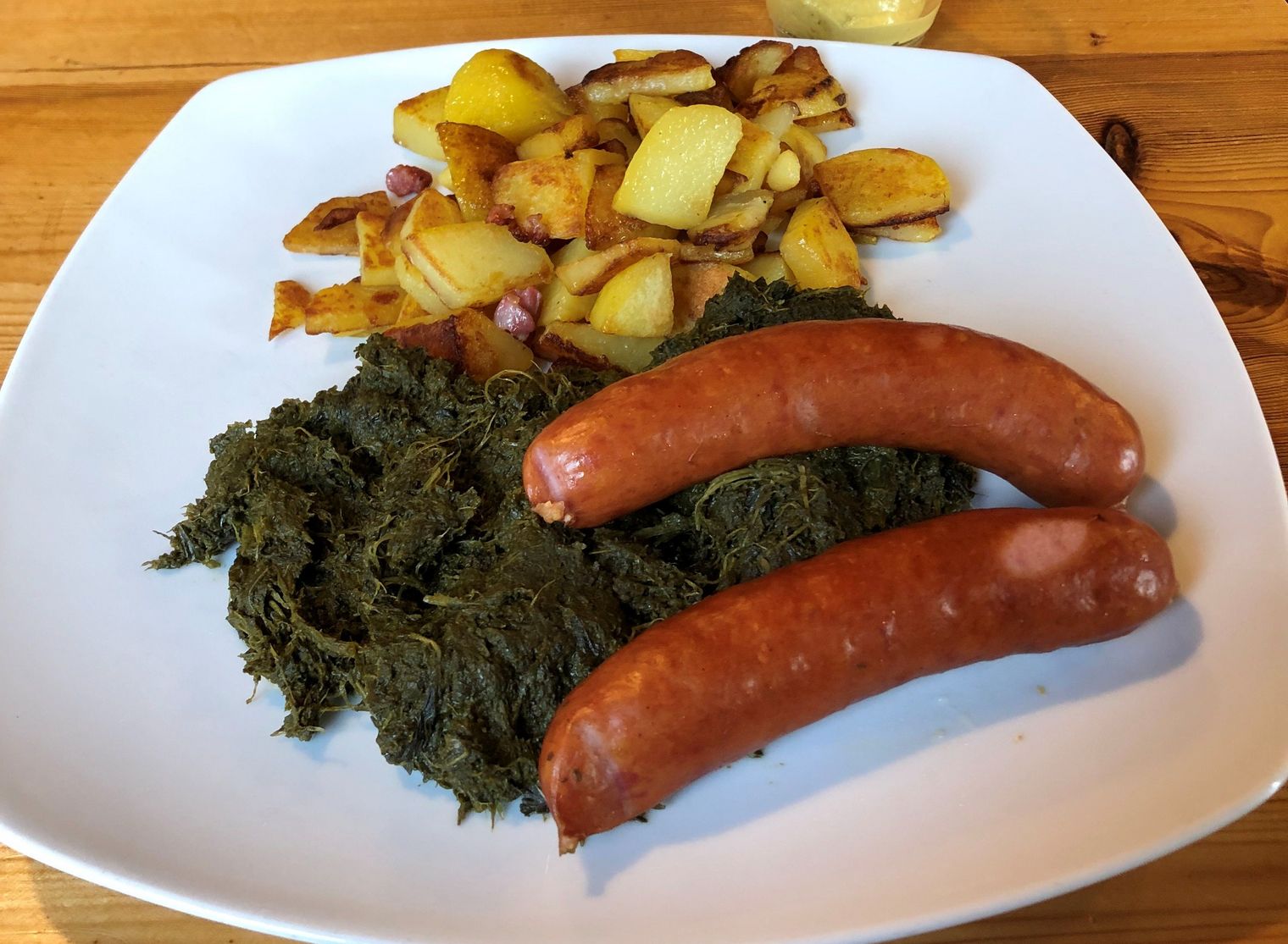 Teller mit Grünkohl, zwei Kochwürsten, Bratkartoffeln und Senf auf einem Holztisch.