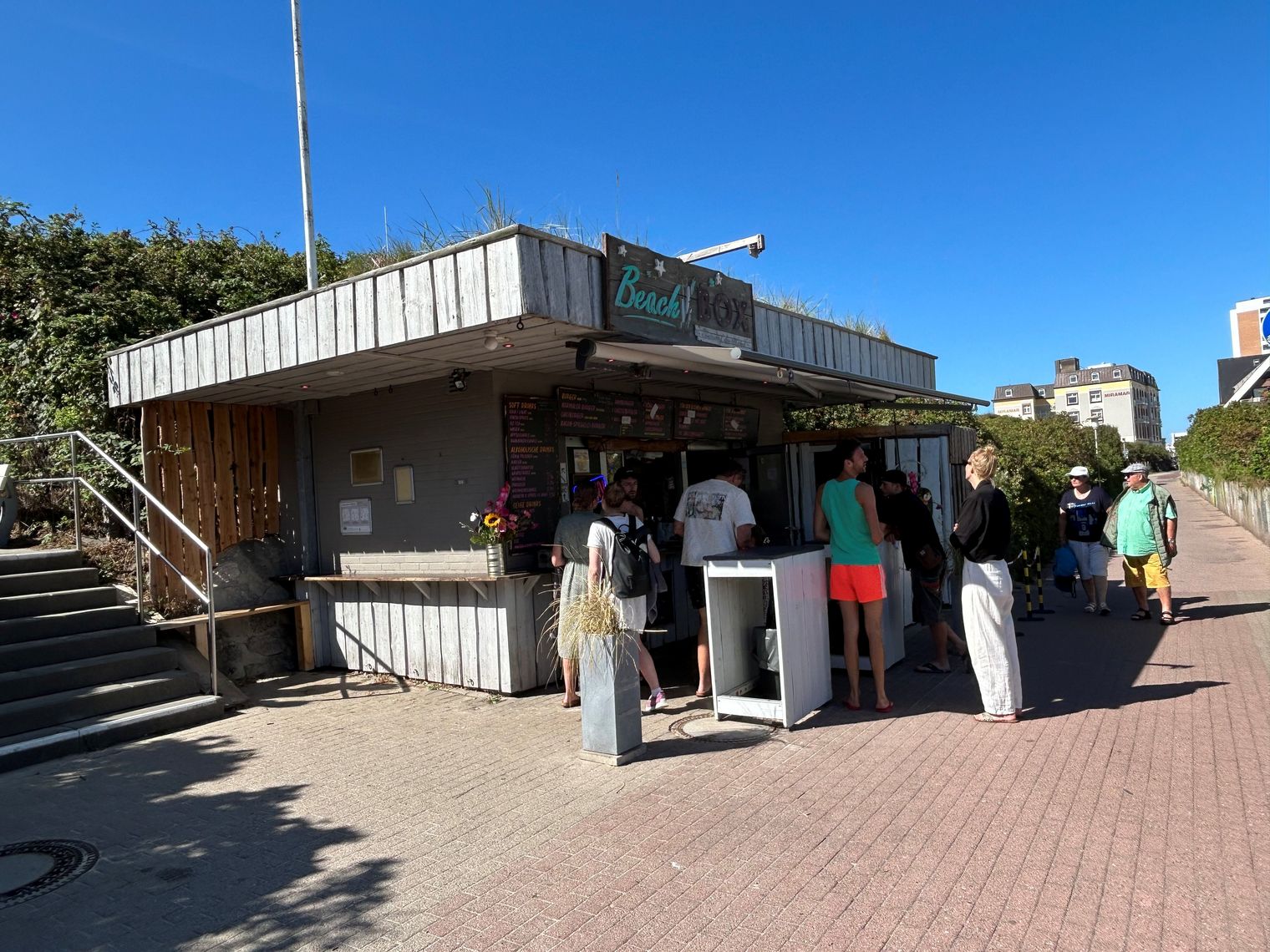 Menschen stehen an einem Strandkiosk mit Holzverkleidung bei strahlendem Sonnenschein.