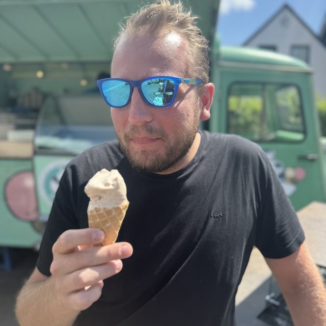 Mann mit blauer Sonnenbrille isst eine Eiswaffel vor einem grünen Eiswagen.