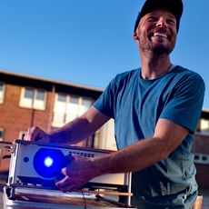 Lachender Mann richtet im Freien einen leuchtenden Projektor ein.
