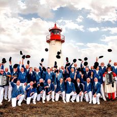 Eine Gruppe eines Shantychors in blauen Jacken und weißen Hosen wirft schwarze Mützen in die Luft vor einem Leuchtturm.