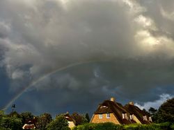 Grauer Himmel mit Regenbogen über angeleuchteten Reetdachhäusern