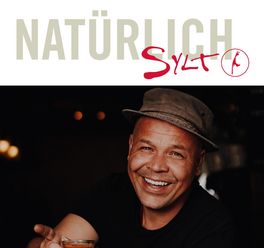  Ein lächelnder Mann mit Hut und Tattoo hält ein Getränk in der Hand.