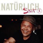  Ein lächelnder Mann mit Hut und Tattoo hält ein Getränk in der Hand.
