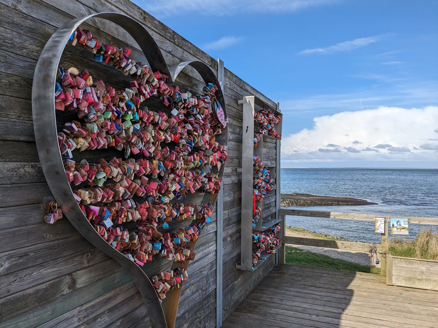Metallherz voller bunter Liebesschlösser an einer Holzwand vor Küstenlandschaft und ruhigem Meer.