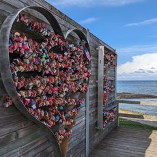 Metallherz voller bunter Liebesschlösser an einer Holzwand vor Küstenlandschaft und ruhigem Meer.