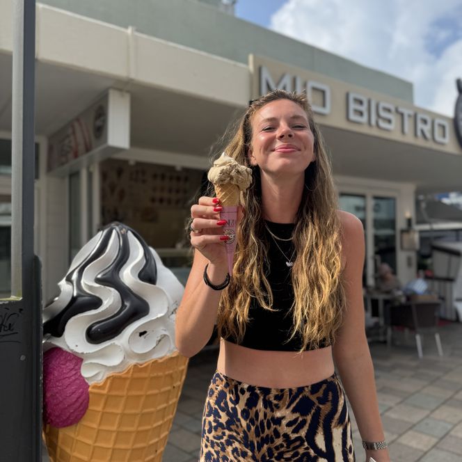 Frau mit langem Haar hält ein Eis vor einem Bistro. Daneben steht eine große Eistüten-Figur.