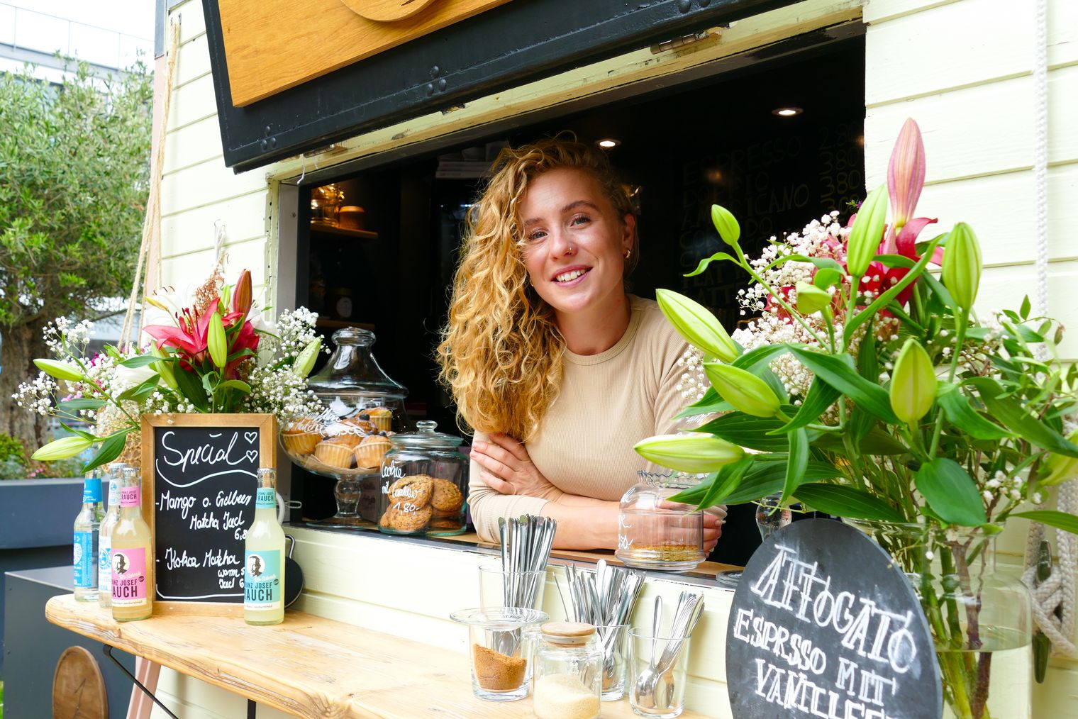 Frau lächelt aus Caféfenster, flankiert von Blumen und Snacks