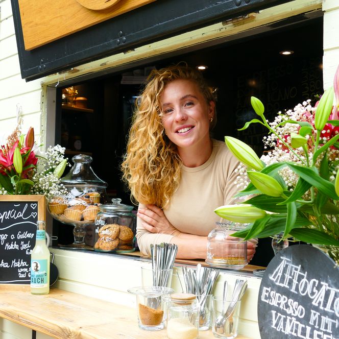 Frau lächelt aus Caféfenster, flankiert von Blumen und Snacks