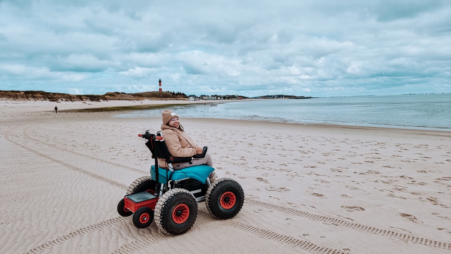 Alternativer Text: Frau fährt im geländegängigen Rollstuhl am Strand entlang.