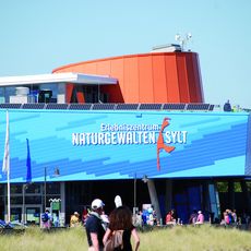 Blaues Gebäude des Erlebniszentrums Naturgewalten Sylt mit Besuchern und Strandgras im Vordergrund.