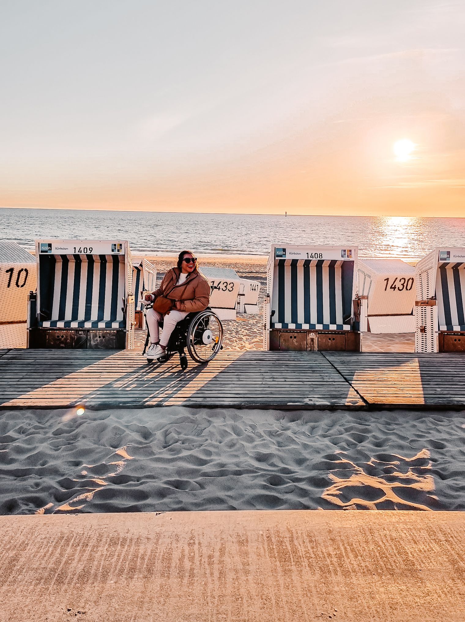 Eine Frau im Rollstuhl sitzt zwischen Strandkörben auf einem barrierefreien Holzsteg am Strand von Westerland, die Sonne geht unter.