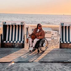 Eine Frau im Rollstuhl sitzt zwischen Strandkörben auf einem barrierefreien Holzsteg am Strand von Westerland, die Sonne geht unter.