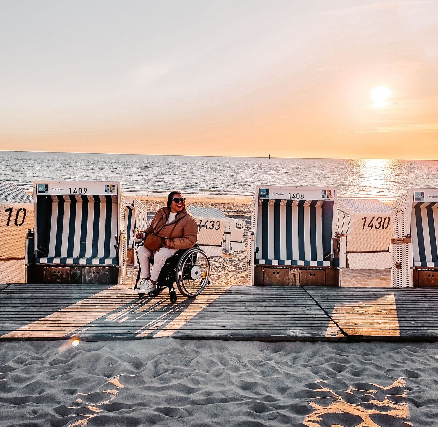 Eine Frau im Rollstuhl sitzt zwischen Strandkörben auf einem barrierefreien Holzsteg am Strand von Westerland, die Sonne geht unter.