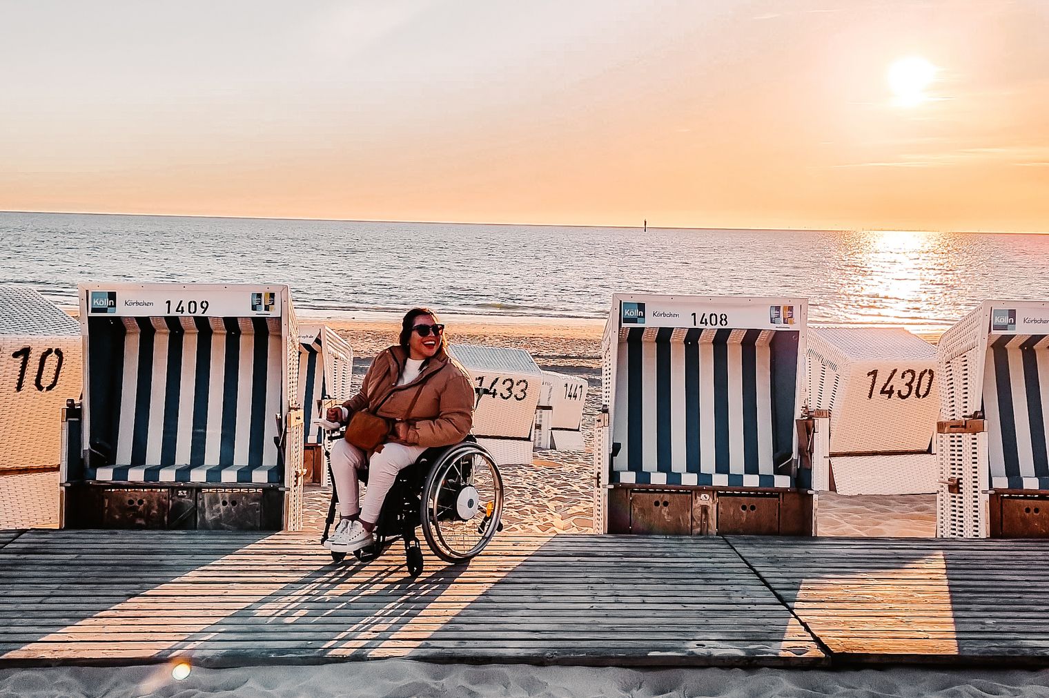 Eine Frau im Rollstuhl sitzt zwischen Strandkörben auf einem barrierefreien Holzsteg am Strand von Westerland, die Sonne geht unter.