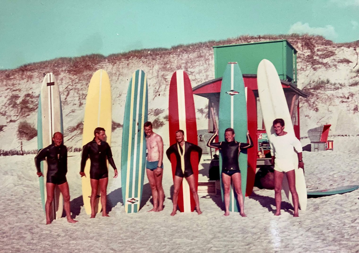 Historisches Foto: Sieben Männer mit bunten Longboards stehen lachend im Sand vor einer Düne auf Sylt.