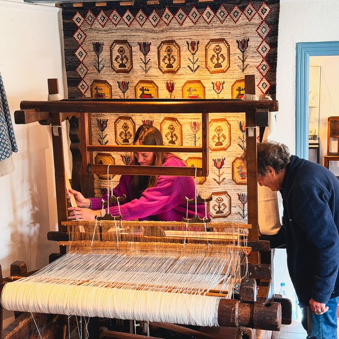 Zwei Personen arbeiten gemeinsam an einem großen Holzwebstuhl.