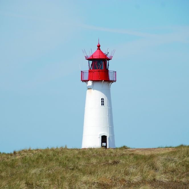 Weiß-roter Leuchtturm in einer Dünenlandschaft unter blauem Himmel.