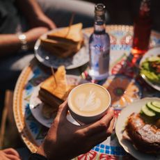 Hand hält Cappuccino mit Latte Art, umgeben von Sandwiches, Croissants und Getränken auf einem bunten Tisch.