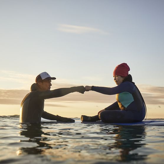 Zwei Surfer auf Boards begrüßen sich mit Faustgruß bei Sonnenuntergang.