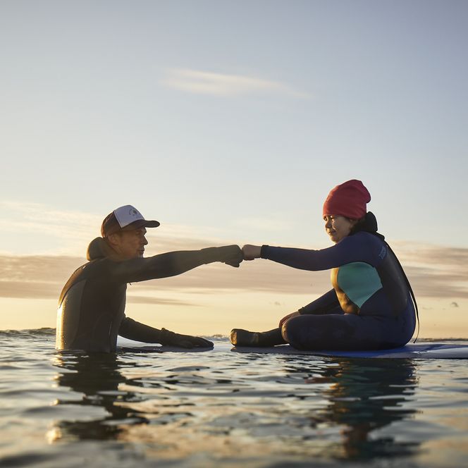 Zwei Surfer auf Boards begrüßen sich mit Faustgruß bei Sonnenuntergang.