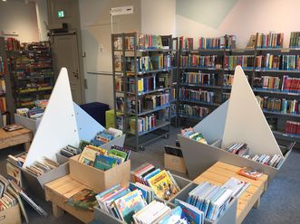 Freundlicher Kinderbuchbereich in einer Bibliothek mit segelbootförmigen Regalen und vielen bunten Büchern.