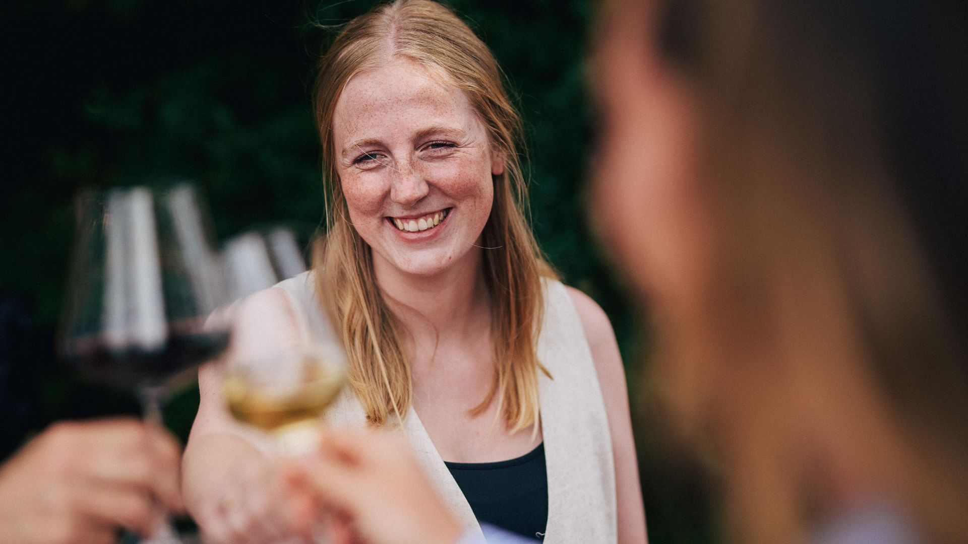 Lächelnde Frau stößt mit anderen Gästen bei einem Glas Wein in einer sommerlichen Umgebung an