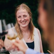 Lächelnde Frau stößt mit anderen Gästen bei einem Glas Wein in einer sommerlichen Umgebung an