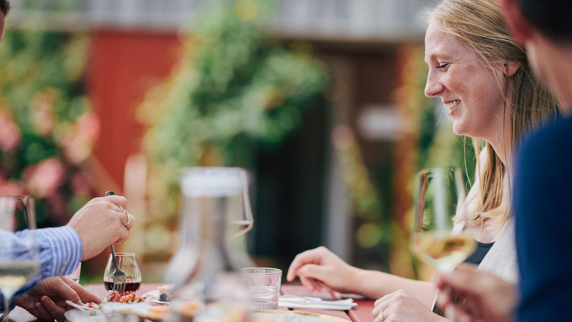 Lächelnde Frau bei einem Essen im Käseklub in Keitum auf Sylt, mit Wein, Wasser und Speisen auf einem Tisch im sommerlichen Ambiente.