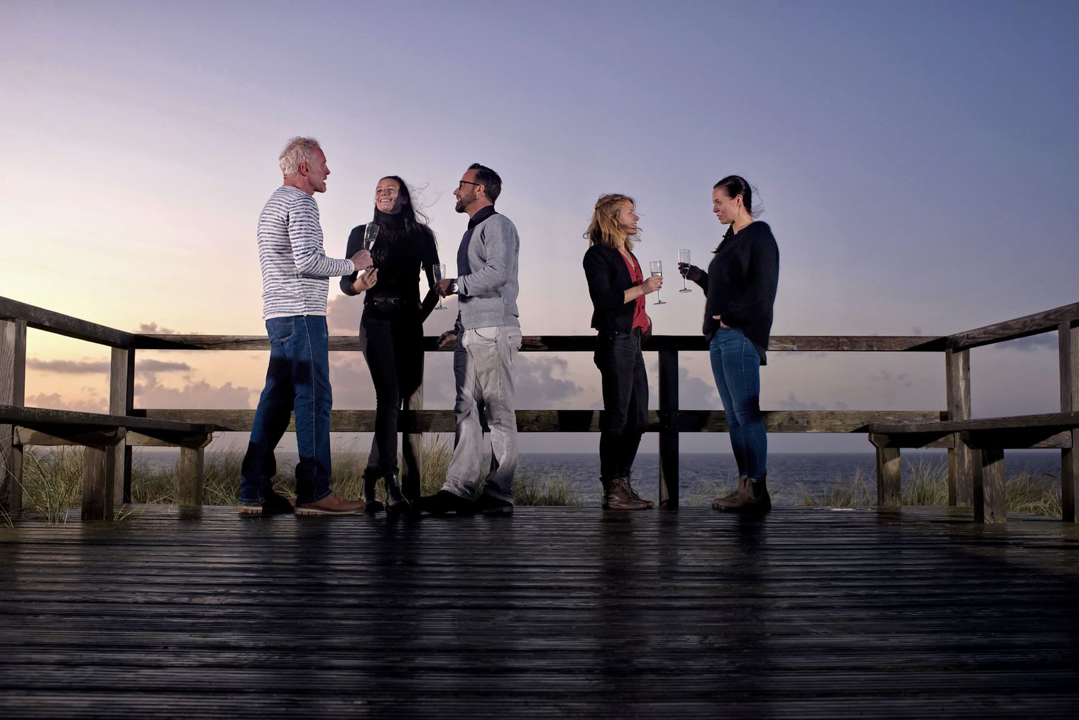 Fünf Personen stehen auf einer hölzernen Plattform am Meer bei Sonnenuntergang und unterhalten sich, während sie Gläser in den Händen halten.