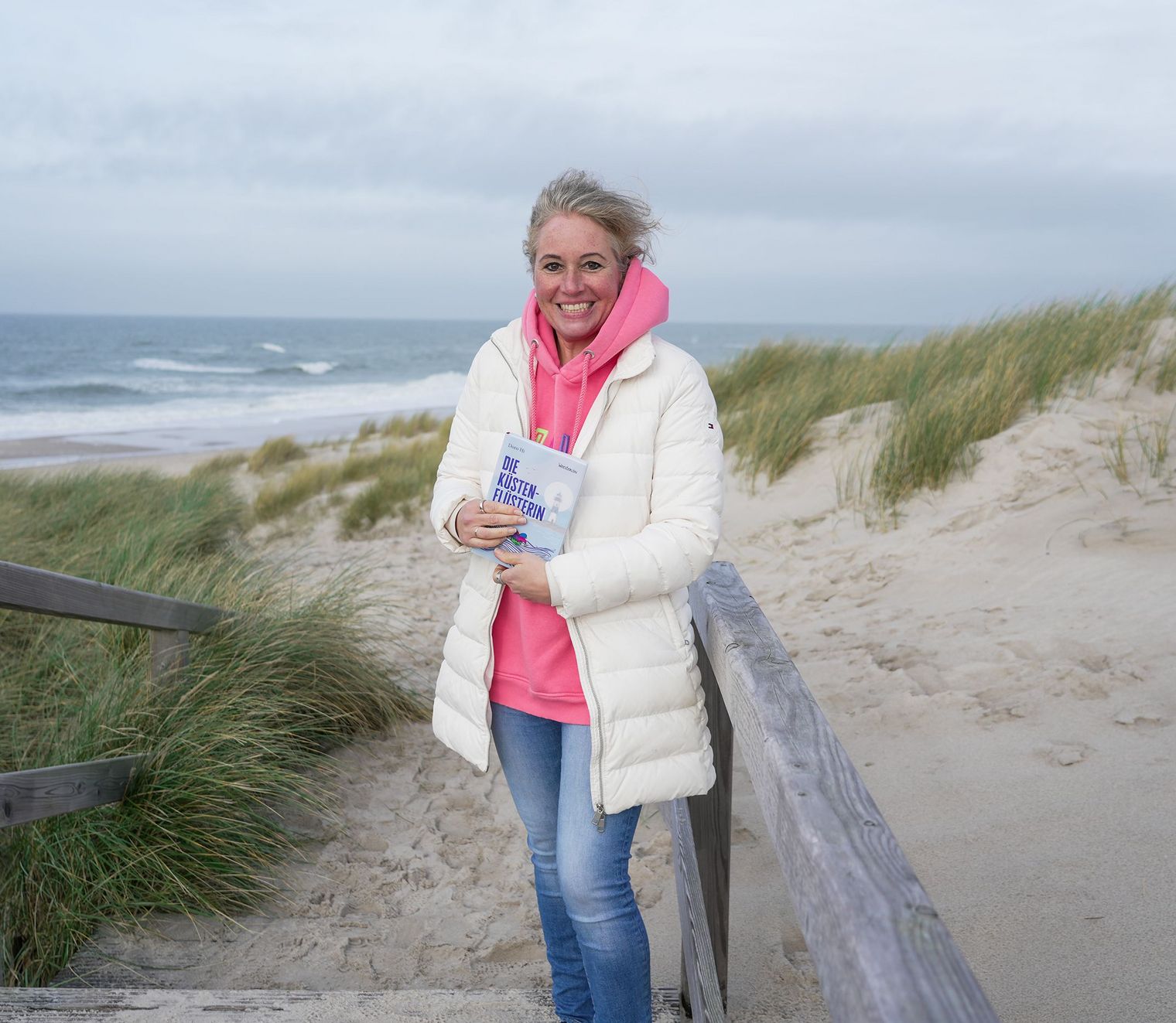 Frau mit Buch steht auf Holzsteg zwischen Dünen und Meer.