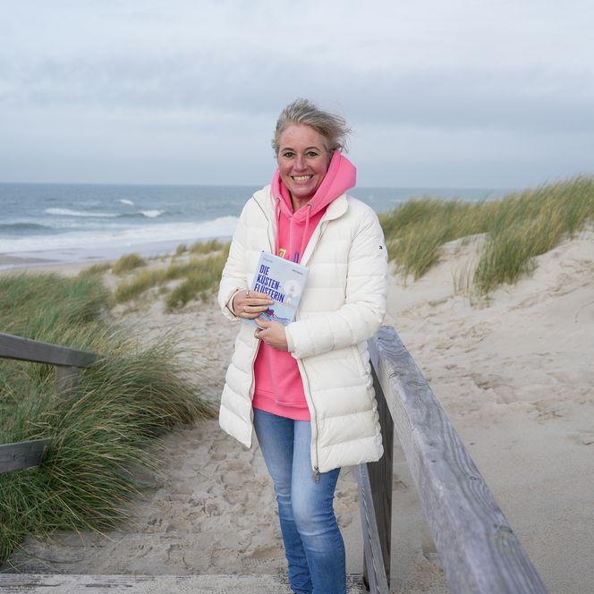 Frau mit Buch steht auf Holzsteg zwischen Dünen und Meer.