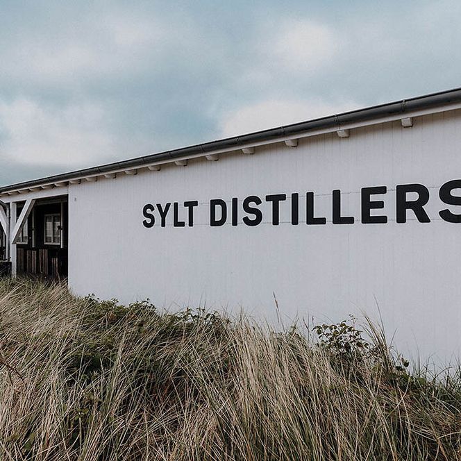 Weißes Gebäude mit Schriftzug „Sylt Distillers“ in Dünenlandschaft