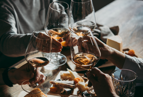 Vier Personen stoßen mit Weißweingläsern über Käseplatte an.