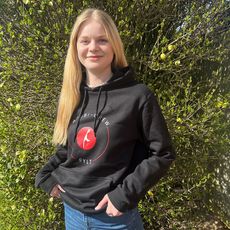 Eine junge Frau mit langen blonden Haaren steht vor einem grünen Busch mit kleinen Knospen. Sie trägt einen schwarzen Kapuzenpullover mit dem Logo der Azubi-Crew Sylt 2024 und hat ihre Hände in den Hosentaschen. Sie lächelt sanft in die Kamera.