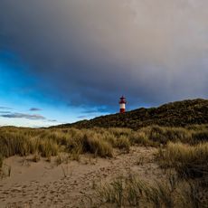 Leuchtturm auf Düne bei dramatischem Himmel mit Sand und Gras im Vordergrund.