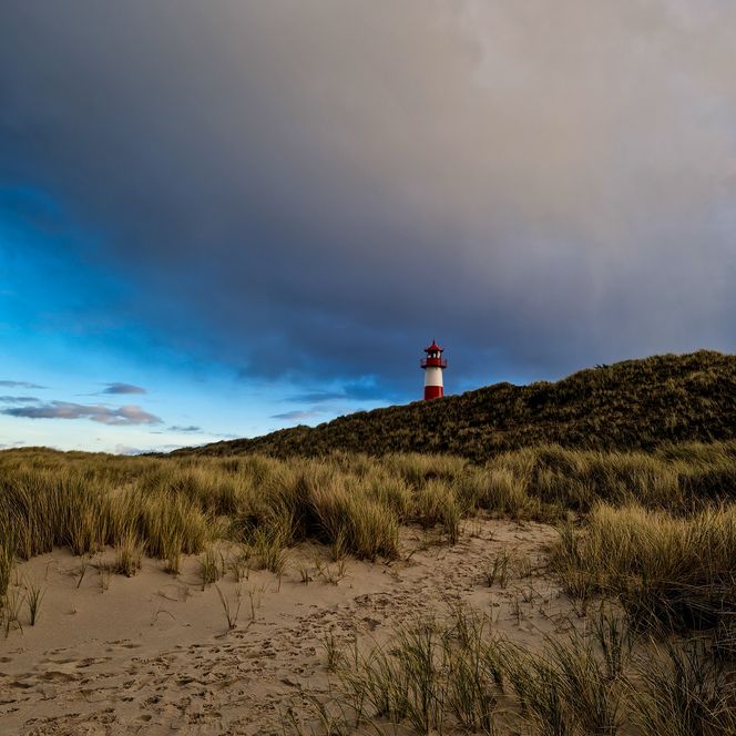 Leuchtturm auf Düne bei dramatischem Himmel mit Sand und Gras im Vordergrund.