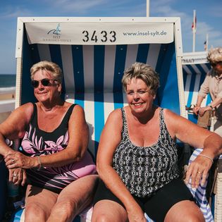 Zwei lächelnde Frauen sitzen bei Sonnenschein in einem blau-weißen Strandkorb an der Strandpromenade mit Meerblick.