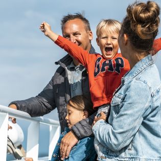 Mehrere Familienmitglieder stehen lachend an der Reling einer Fähre unter blauem Himmel.
