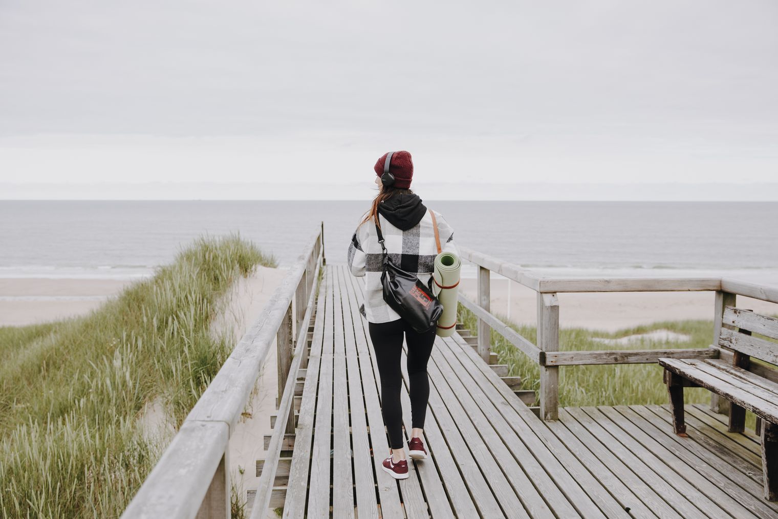 Eine Frau mit Kopfhörern und Yogamatte geht über einen Holzbohlenweg. Vor ihr liegt der Strand.