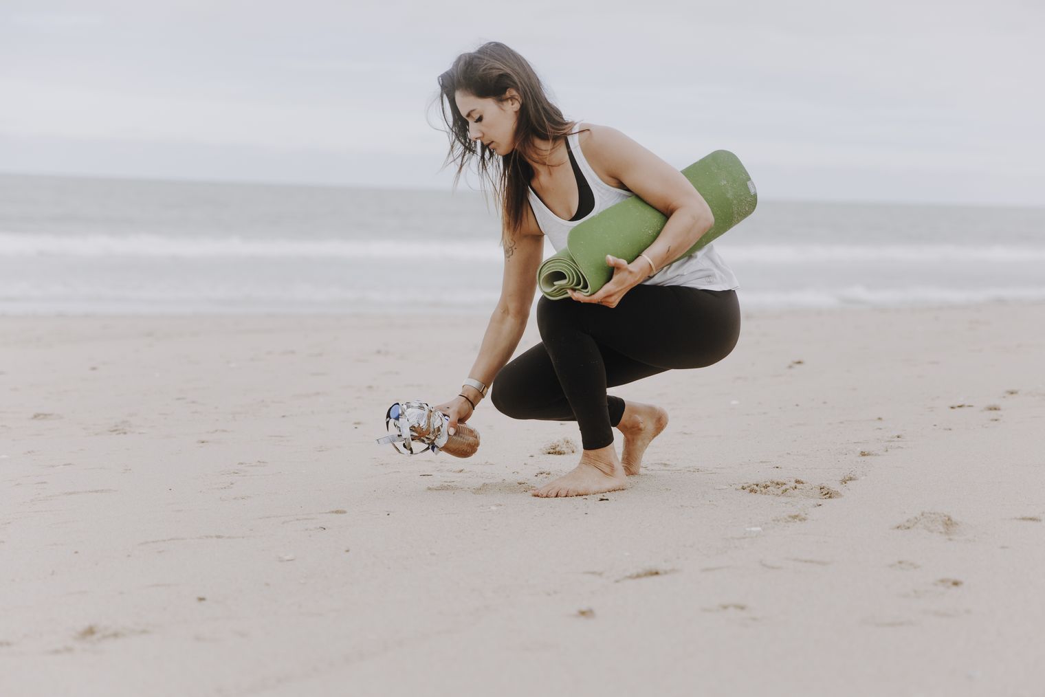 Eine Frau sammelt achtsam Müll am Strand ein, während sie eine zusammengerollte Yogamatte unter dem Arm trägt.