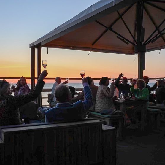  Menschen stoßen mit Weingläsern auf einer Terrasse bei Sonnenuntergang mit Meerblick an.