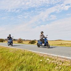 Zwei Personen fahren mit E-Rollern auf einer Landstraße auf Sylt