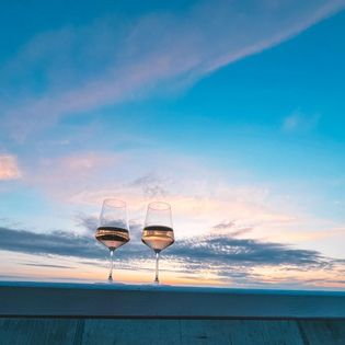 Zwei Weingläser auf einer Holzbalustrade vor einem farbenfrohen Sonnenuntergang mit blauem Himmel und Wolken.