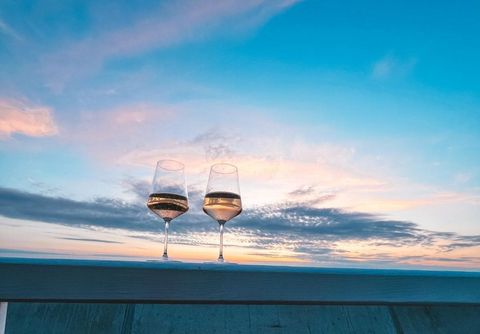 Due bicchieri di vino su una balaustra di legno di fronte a un tramonto colorato con cielo azzurro e nuvole.