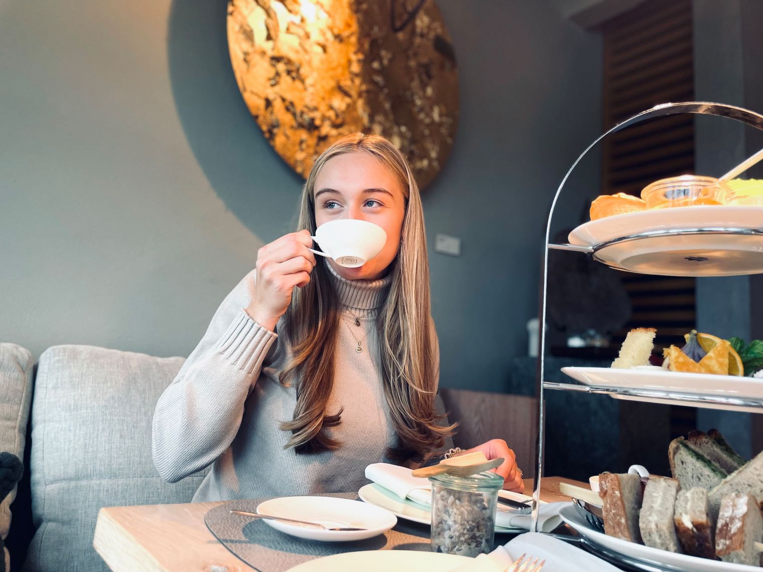 Eine junge Frau genießt eine Tasse Tee in einem gemütlichen Café, vor sich eine Etagere mit Gebäck und Sandwiches.