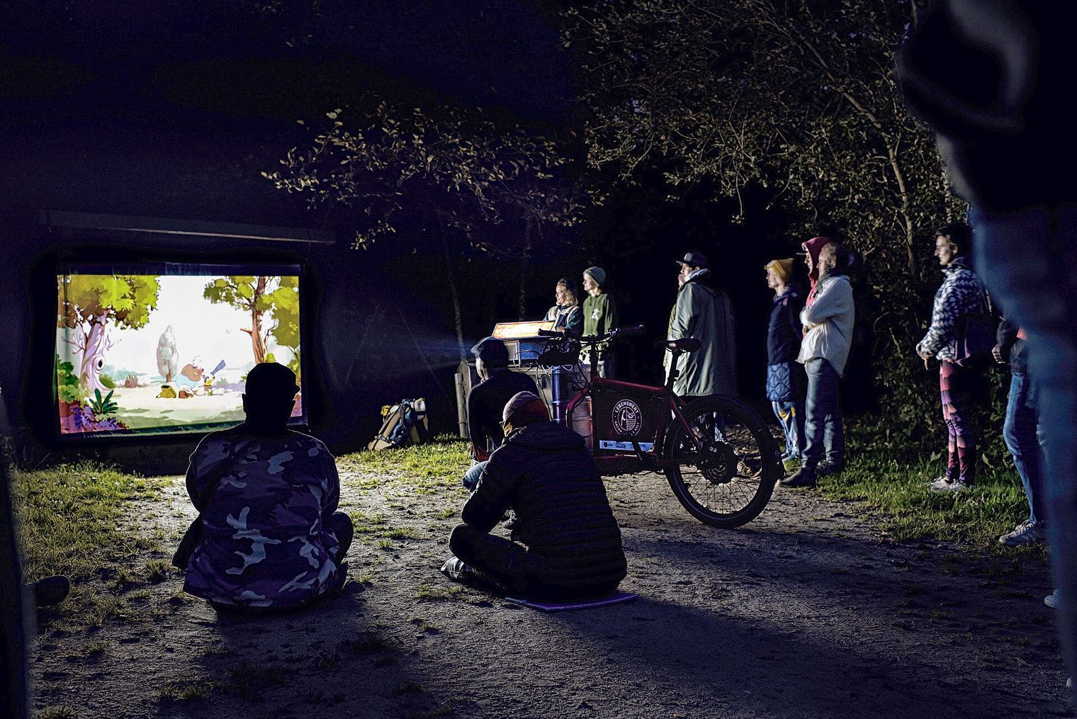 Menschen sehen nachts im Wald einen Animationsfilm auf Leinwand.