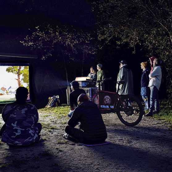 Menschen sehen nachts im Wald einen Animationsfilm auf Leinwand.