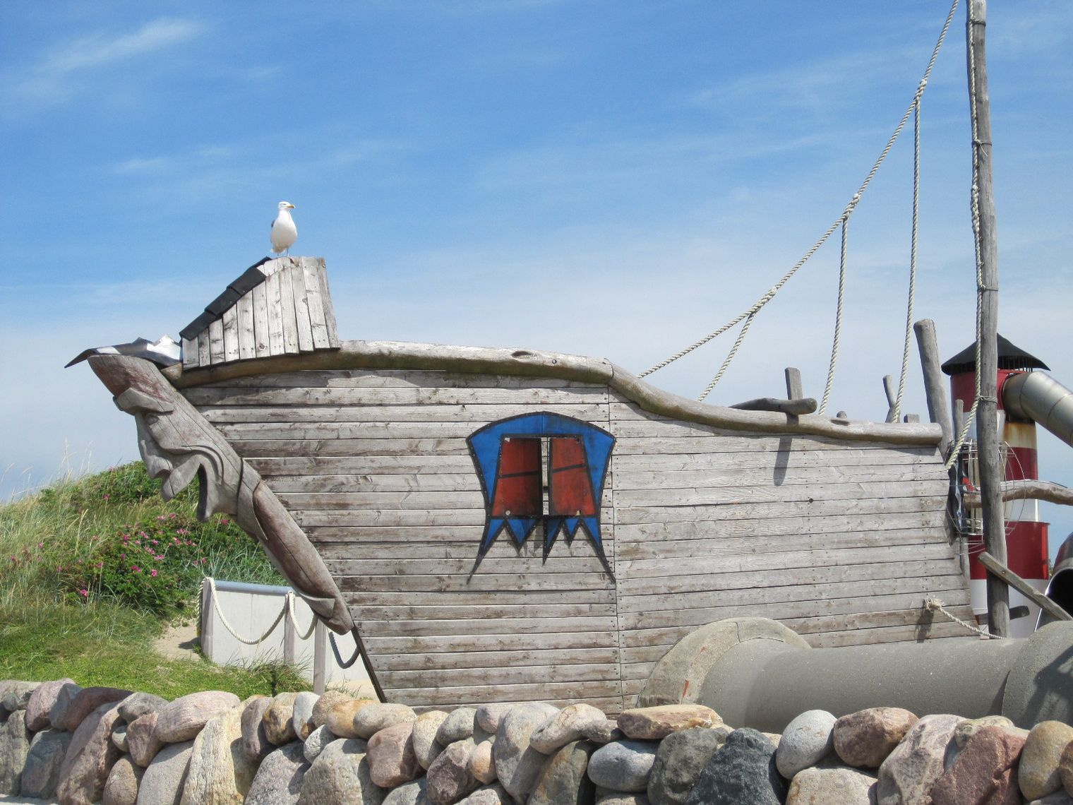 Ein Piratenspielschiff aus Holz mit geschnitztem Bug und rotem Fenster steht auf einem Kinderspielplatz unter blauem Himmel.