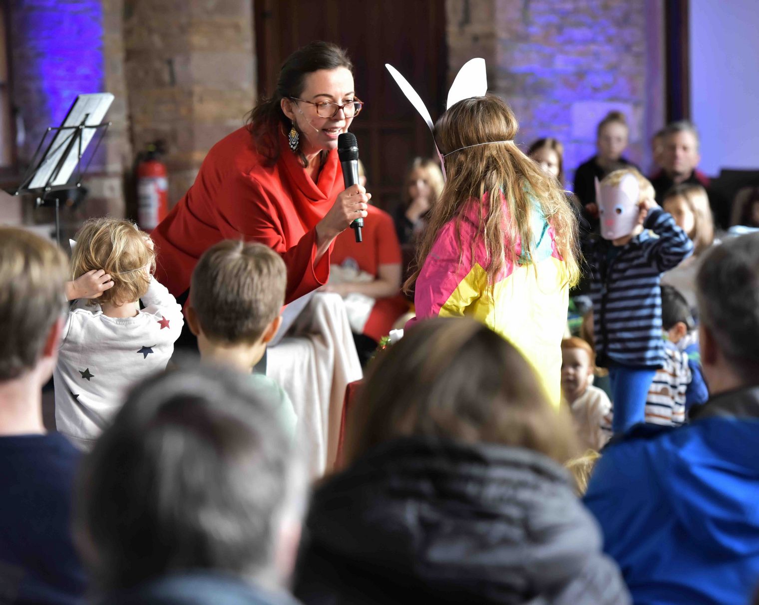 Moderatorin spricht mit einem Kind in Hasenmaske während eines Konzerts, Kinder im Publikum interagieren.